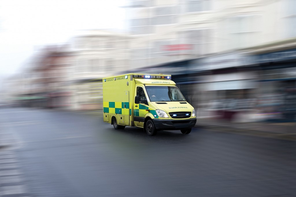 Picture of a yellow emergency ambulance rushing through a city to the hospital with full flashing blue lights and siren. Speed is emphasized by the motion blurred urban background. Vehicle is generic: no visible making or brand. License plate and other markings are also removed.