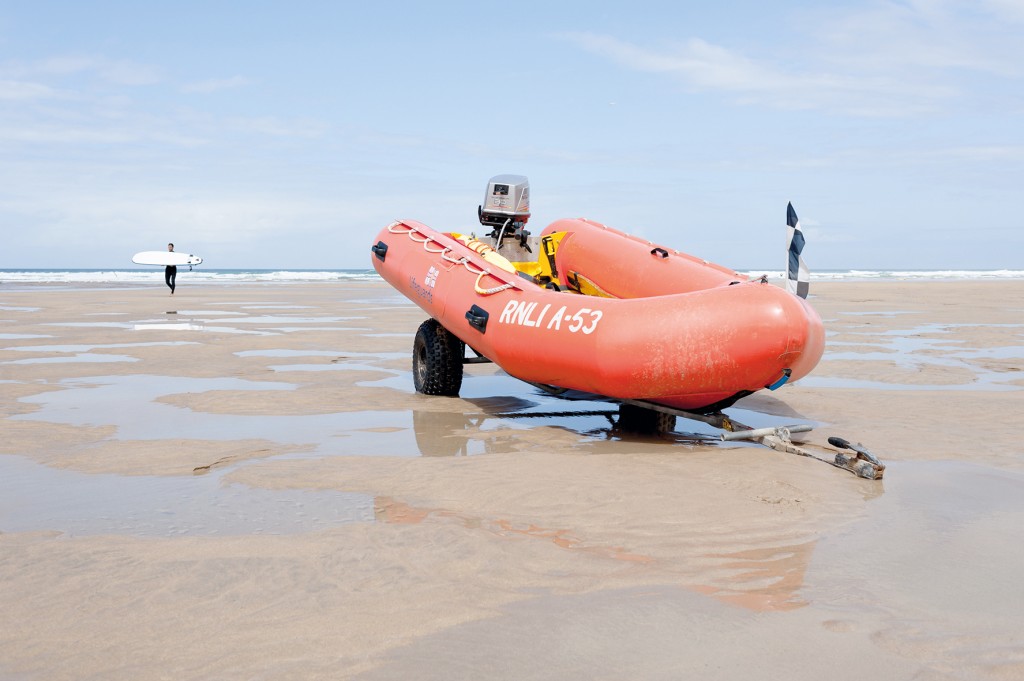 An RNLI inflatable lifeboat resting on a trailer on a Cornish beach with a surfer carrying his surf board in the background.